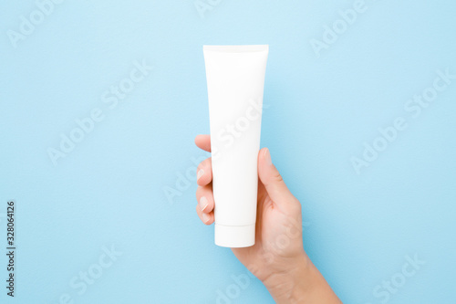 Young woman hand holding and showing white tube on light blue background. Pastel color. Care about clean and soft body skin. Daily beauty product. Closeup. Top down view.