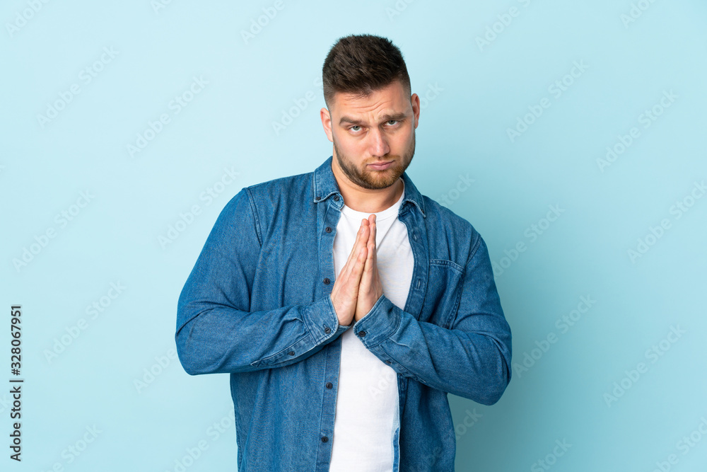 Russian handsome man isolated on blue background keeps palm together. Person asks for something Russian handsome man isolated on blue background keeps palm together. Person asks for something