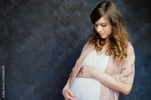 Expectant mom touching her belly. Beautiful pregnant young woman on dark background. Copy space.