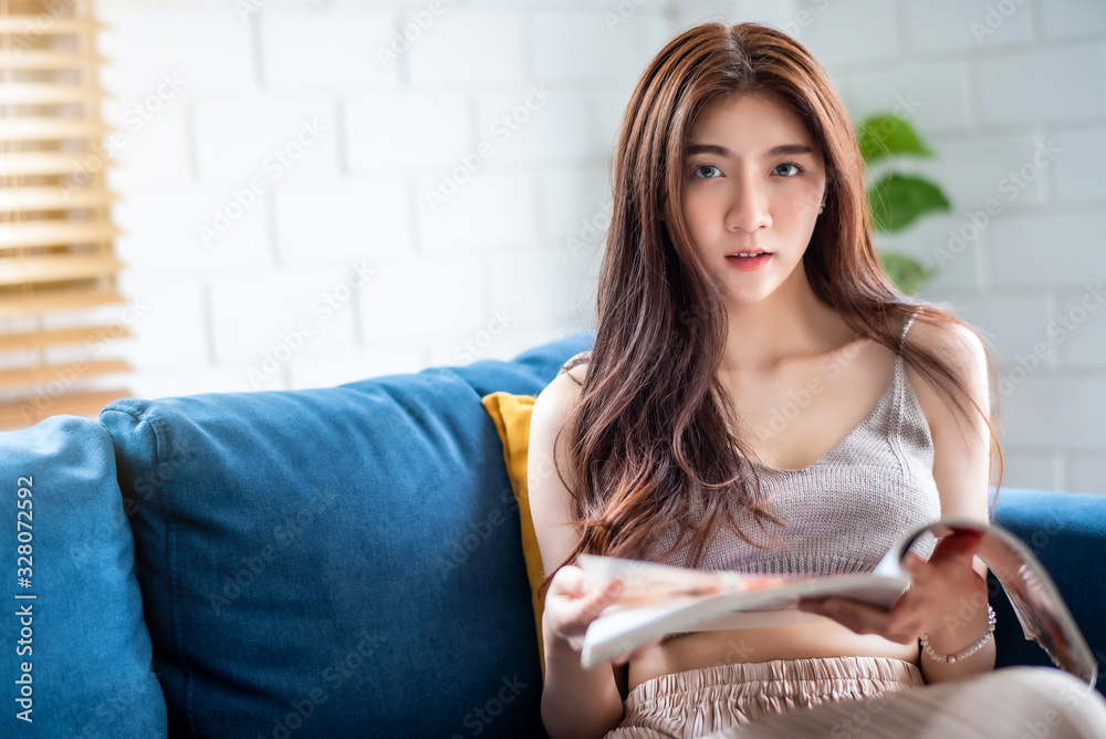 Young beautiful Asian woman sitting on blue sofa and reading magazine at living room in the morning Young beautiful Asian woman sitting on blue sofa and reading magazine at living room in the morning
