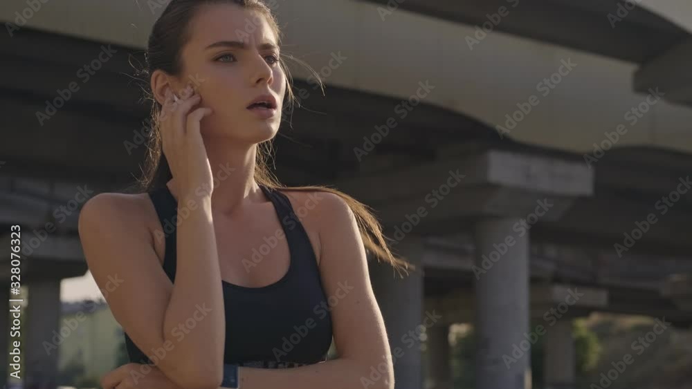 An exhausted slim young woman is using her tracker and earphones while running outdoors near the bridge in the city An exhausted slim young woman is using her tracker and earphones while running outdoors near the bridge in the city