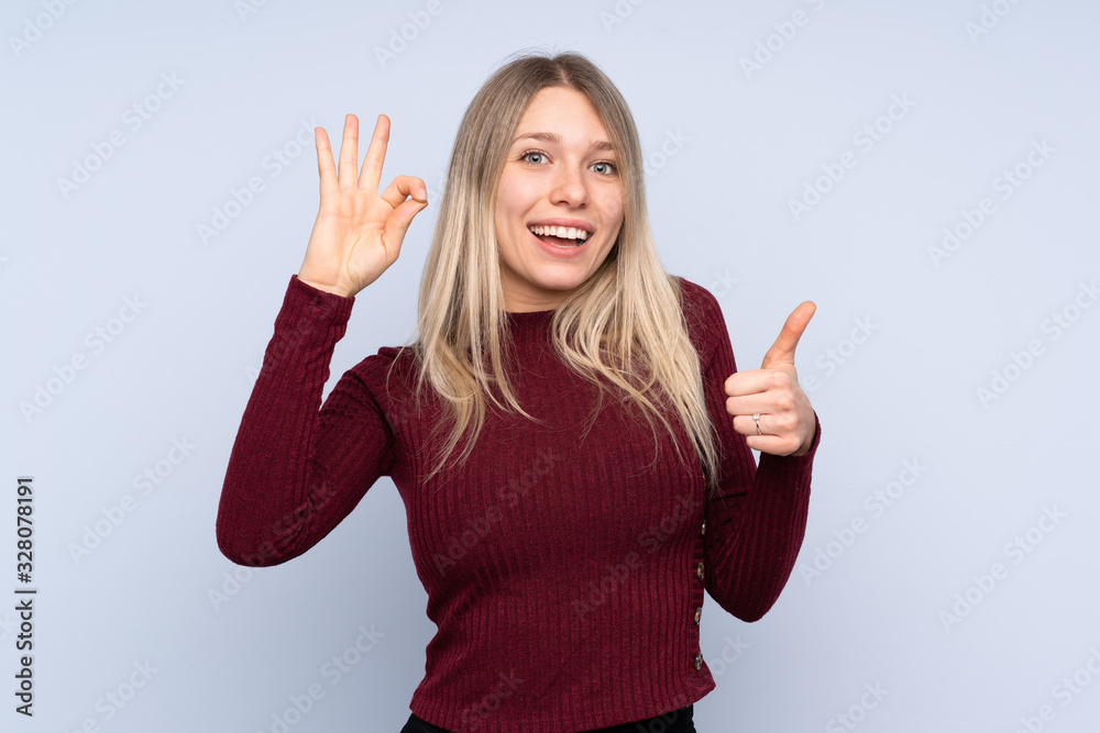 Young blonde woman over isolated blue background showing ok sign and thumb up gesture Young blonde woman over isolated blue background showing ok sign and thumb up gesture