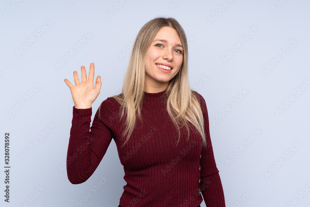Young blonde woman over isolated blue background saluting with hand with happy expression Young blonde woman over isolated blue background saluting with hand with happy expression