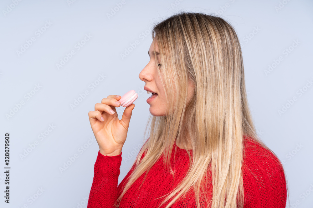 Young blonde woman over isolated blue background holding colorful French macarons and eating it Young blonde woman over isolated blue background holding colorful French macarons and eating it