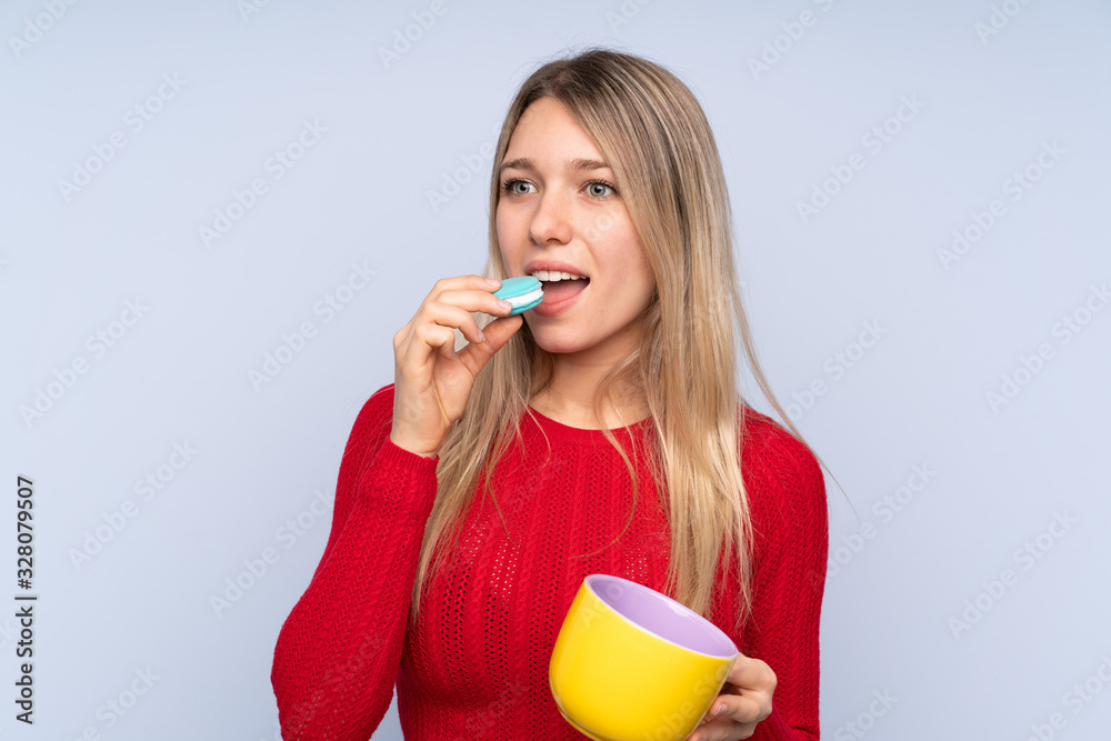Young blonde woman over isolated blue background holding colorful French macarons and a cup of milk