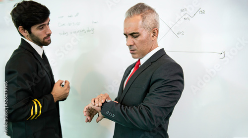 Confident businessman look at his wristwatch while discussion with young pilot man and writing graph on whiteboard about aviation businessn,startup,business team,economics,analytics and statistics. photo