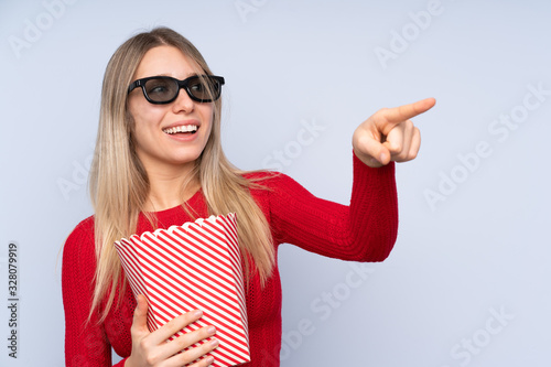 Young blonde woman over isolated blue background with 3d glasses and holding a big bucket of popcorns while pointing away