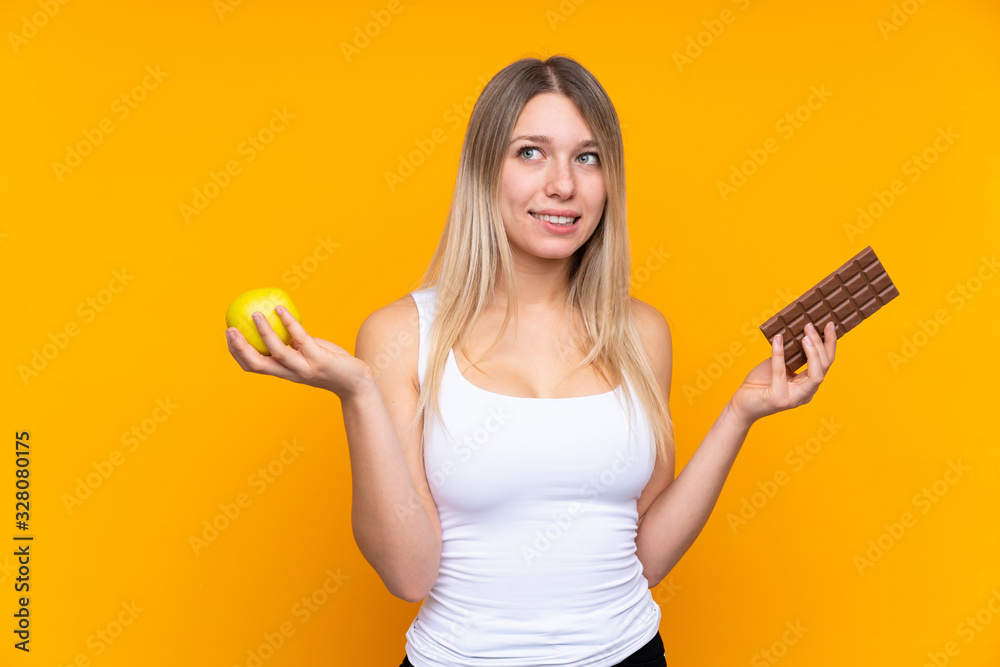 Young blonde woman over isolated blue background having doubts while taking a chocolate tablet in one hand and an apple in the other Young blonde woman over isolated blue background having doubts while taking a chocolate tablet in one hand and an apple in the other