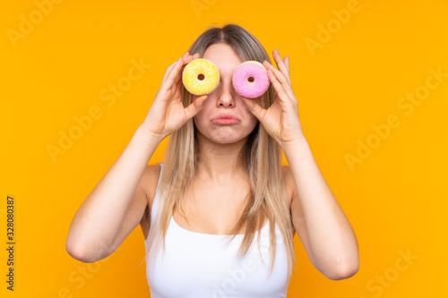 Young blonde woman over isolated blue background holding donuts in eyes with sad expression © luismolinero
