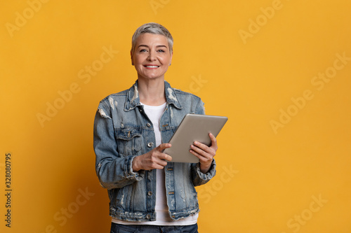 Smiling mature woman with digital tablet in hands on yellow background