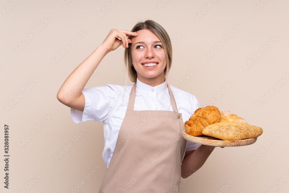 Young blonde girl with apron. Female baker holding a table with several breads having doubts and with confuse face expression Young blonde girl with apron. Female baker holding a table with several breads having doubts and with confuse face expression