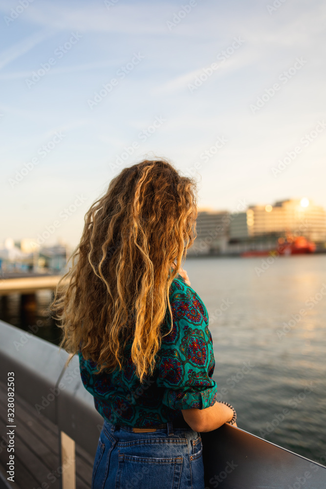 Blond girl with curly hair looking at the sunset by the sea in Barcelona, Catalonia, Spain.