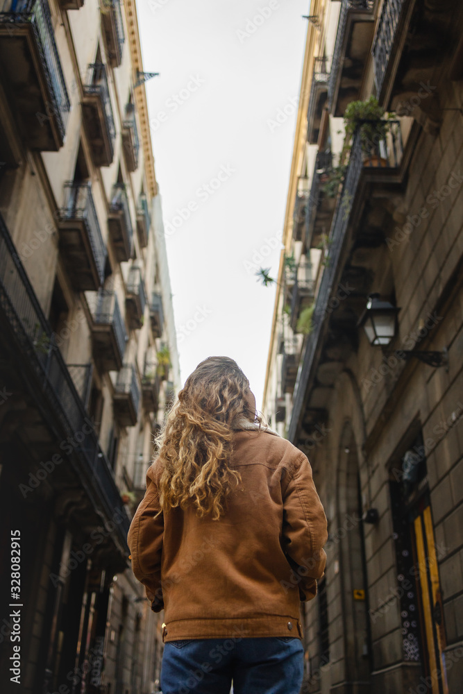 Naklejka premium Blond girl walking through the streets of the center of the city of Barcelona in Catalonia, Spain wearing a jacket and blue jeans. Looking up to the old buldings.