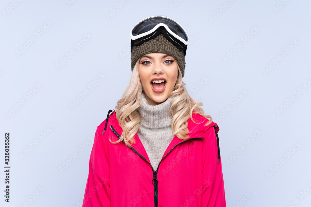 Skier teenager girl with snowboarding glasses over isolated blue background with surprise facial expression