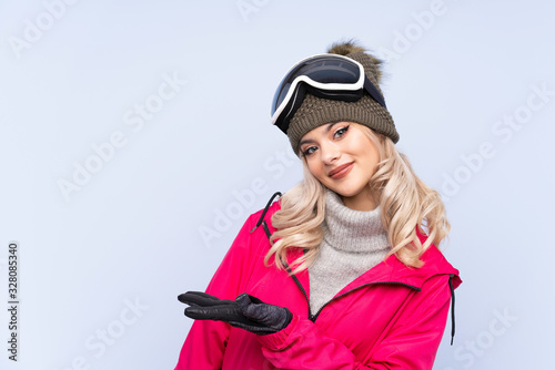 Skier teenager girl with snowboarding glasses over isolated blue background extending hands to the side for inviting to come © luismolinero