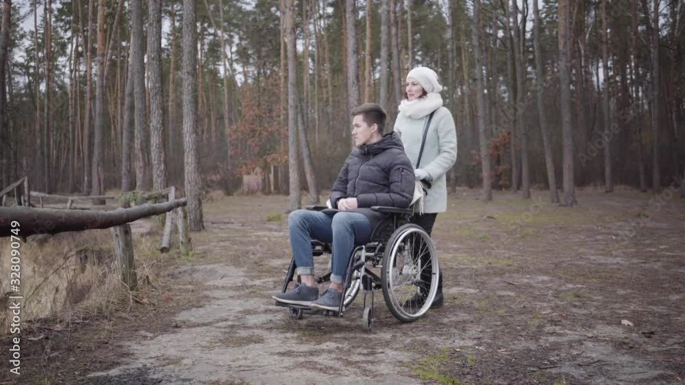 Portrait of Caucasian woman talking with young disabled man in wheelchair. Invalid tender spending time with patient in autumn forest. Disability, lifestyle, help, care. Portrait of Caucasian woman talking with young disabled man in wheelchair. Invalid tender spending time with patient in autumn forest. Disability, lifestyle, help, care.