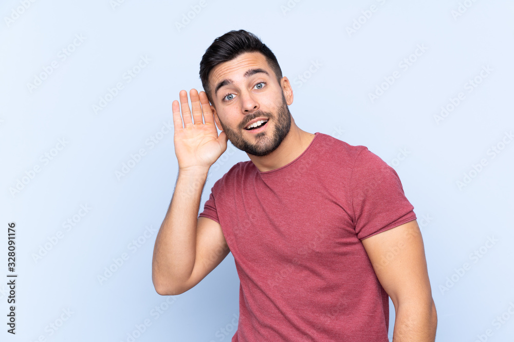 Young handsome man with beard over isolated blue background listening to something by putting hand on the ear