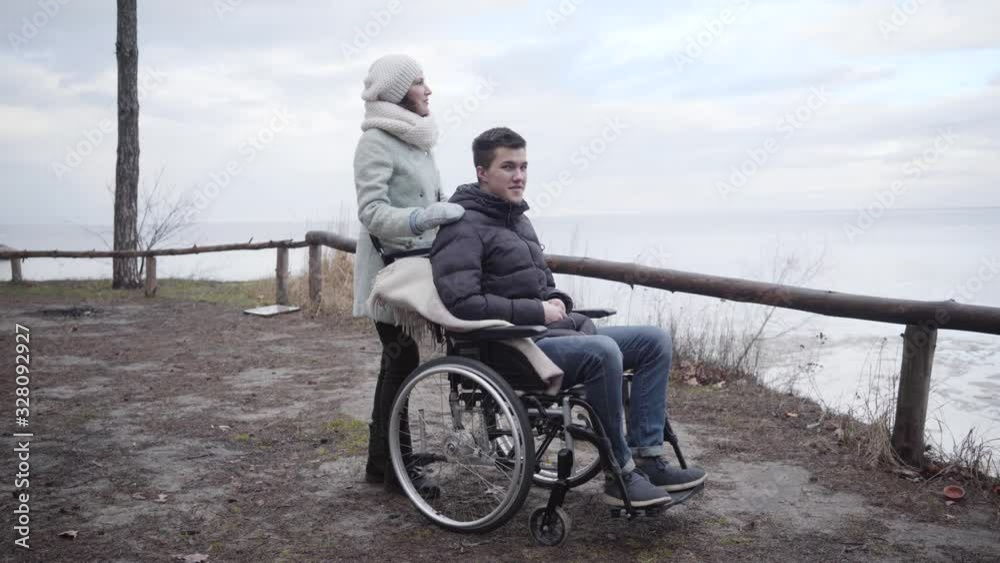Portrait of young positive Caucasian woman touching shoulder of disabled man in wheelchair and pointing away. Strong mother or invalid tender taking care of handicapped boy. Leisure outdoors. Portrait of young positive Caucasian woman touching shoulder of disabled man in wheelchair and pointing away. Strong mother or invalid tender taking care of handicapped boy. Leisure outdoors.
