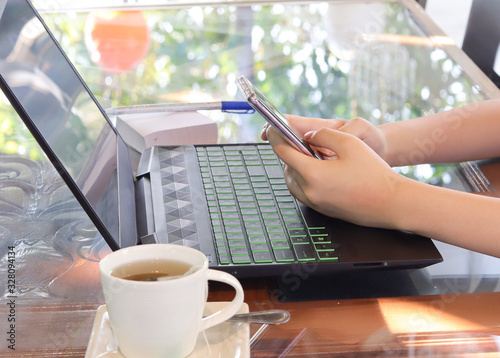 hands holding cell phone and notebook computer on table and top of table is glass and there is tea cup on left hand side.