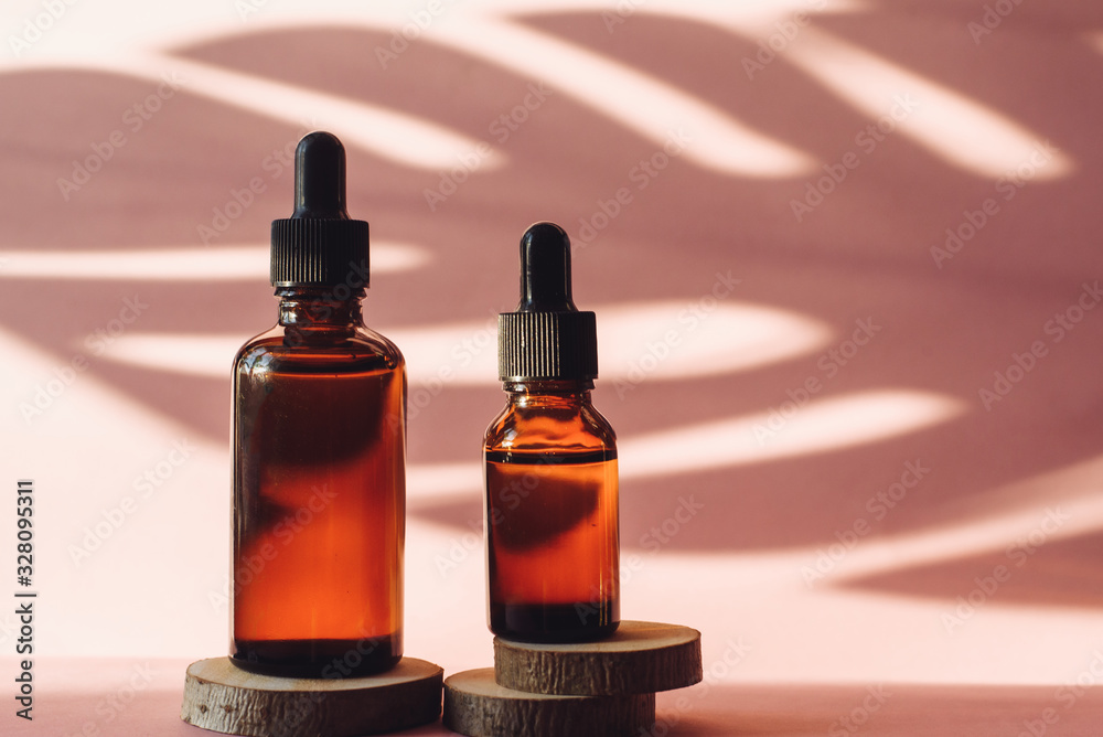 Fototapeta premium Two glass bottles with moisturizing serum on wooden saw cuts on a pink background. Brown tubes with a dropper on the background of a tropical shadow of a palm leaf. The concept of skin care