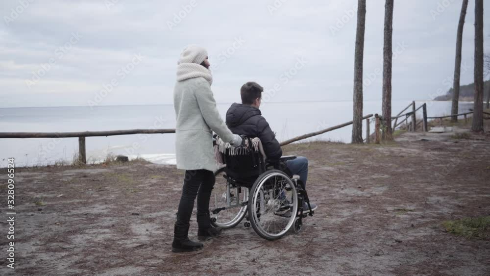 Back side view of woman rolling wheelchair with young Caucasian man along autumn river bank. Invalid tender or mother taking care of handicapped boy outdoors. Leisure, lifestyle, disability. Back side view of woman rolling wheelchair with young Caucasian man along autumn river bank. Invalid tender or mother taking care of handicapped boy outdoors. Leisure, lifestyle, disability.