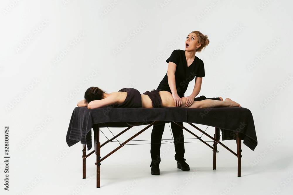 Funny fat burning massage. Portrait of cheerful woman masseur in uniforme near massage table with woman patient in wellness center. White background isolated Funny fat burning massage. Portrait of cheerful woman masseur in uniforme near massage table with woman patient in wellness center. White background isolated