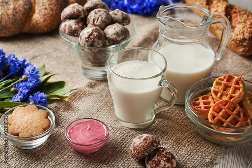 A glass mug with milk and a jug of milk stand on a burlap. Around are waffles  cookies  raspberry honey  gingerbread cookies  bread  and a bouquet of cornflowers