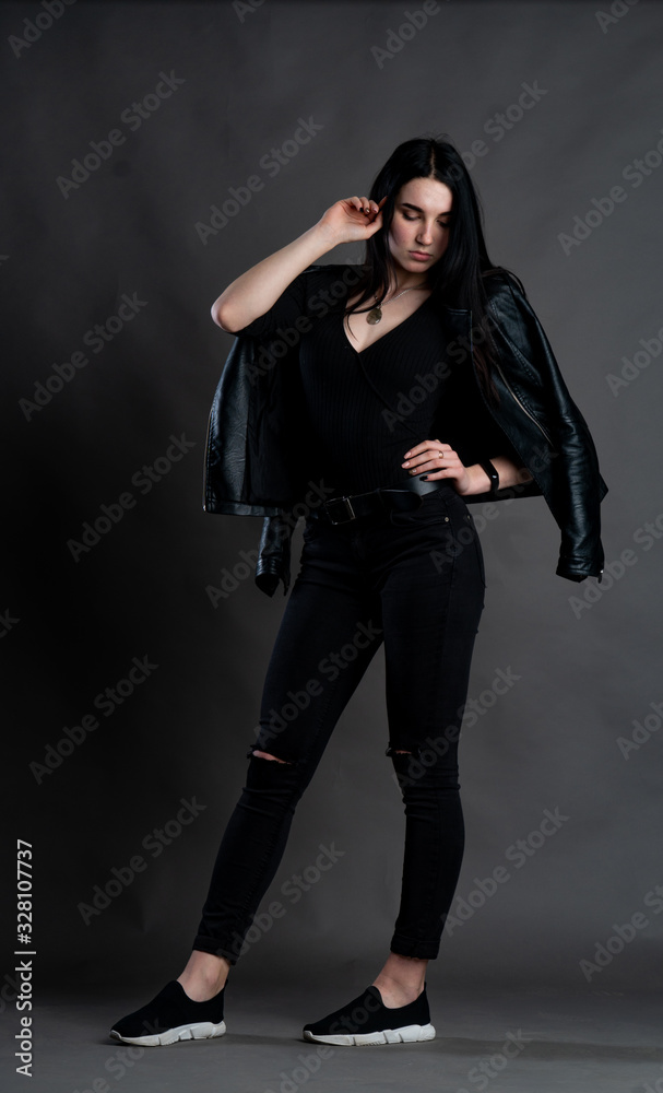 Portrait of young brunette woman sitting on high wooden chair with crossed legs. Fashionable stylish girl wearing wide leg pants. Studio shoot on gray background