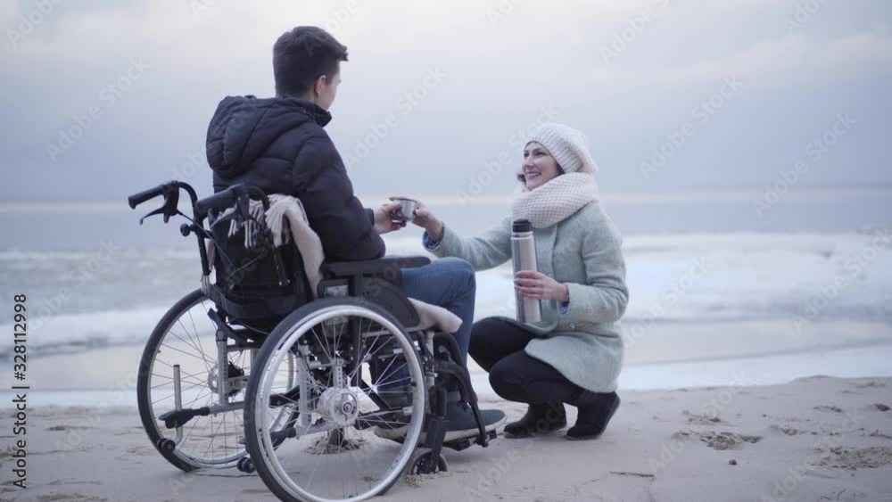Young crippled Caucasian boy in wheelchair drinking tea on sea shore and talking with beautiful woman sitting on hunkers. Disabled young man spending autumn day with positive invalid tender outdoors. Young crippled Caucasian boy in wheelchair drinking tea on sea shore and talking with beautiful woman sitting on hunkers. Disabled young man spending autumn day with positive invalid tender outdoors.