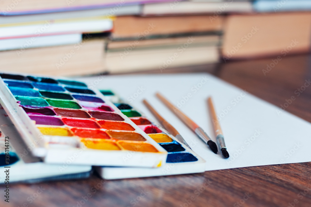 Watercolor paint, brushes well used and sheet of paper on wooden table with stack of books background. Watercolor paint, brushes well used and sheet of paper on wooden table with stack of books background.