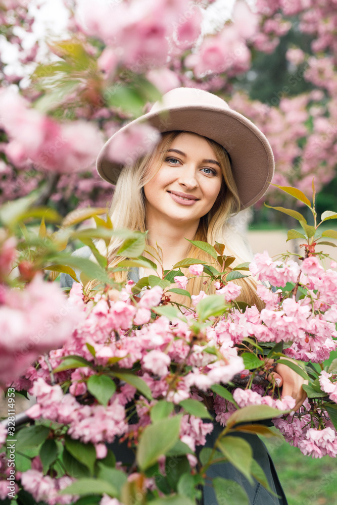 young beautiful blonde girl in a white dress, a gray cloak and a pink hat stands in a park near sakura trees young beautiful blonde girl in a white dress, a gray cloak and a pink hat stands in a park near sakura trees
