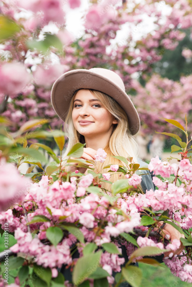 young beautiful blonde girl in a white dress, a gray cloak and a pink hat stands in a park near sakura trees