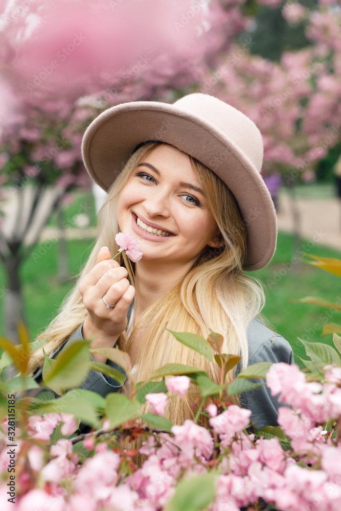 young beautiful blonde girl in a white dress, a gray cloak and a pink hat stands in a park near sakura trees
