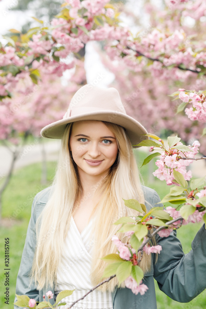young beautiful blonde girl in a white dress, a gray cloak and a pink hat stands in a park near sakura trees young beautiful blonde girl in a white dress, a gray cloak and a pink hat stands in a park near sakura trees