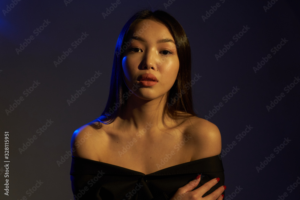 Fototapeta premium Portrait of a young beautiful Asian girl looking directly at the camera. The woman's hair and shoulders in colorful bright UV blue lights. On black background. 