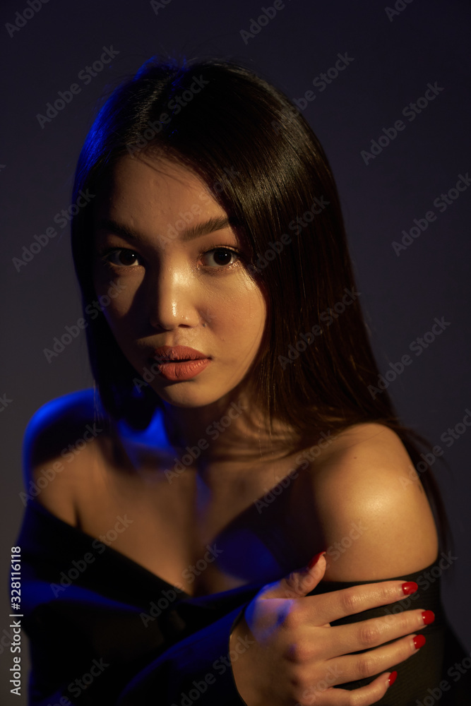 Fototapeta premium Portrait of a young beautiful Asian girl looking directly at the camera. The woman's hair and shoulders in colorful bright UV blue lights. On black background. 