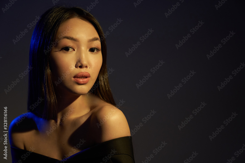 Fototapeta premium Portrait of a young beautiful Asian girl looking directly at the camera. The woman's hair and shoulders in colorful bright UV blue lights. On black background.