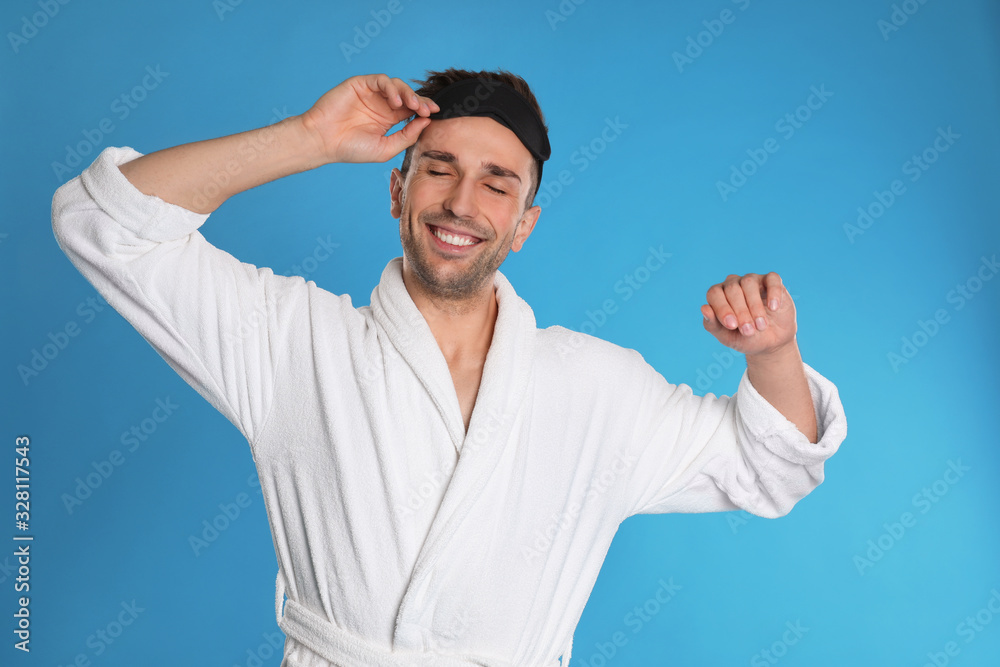 Naklejka premium Happy young man in bathrobe and eye sleeping mask on light blue background
