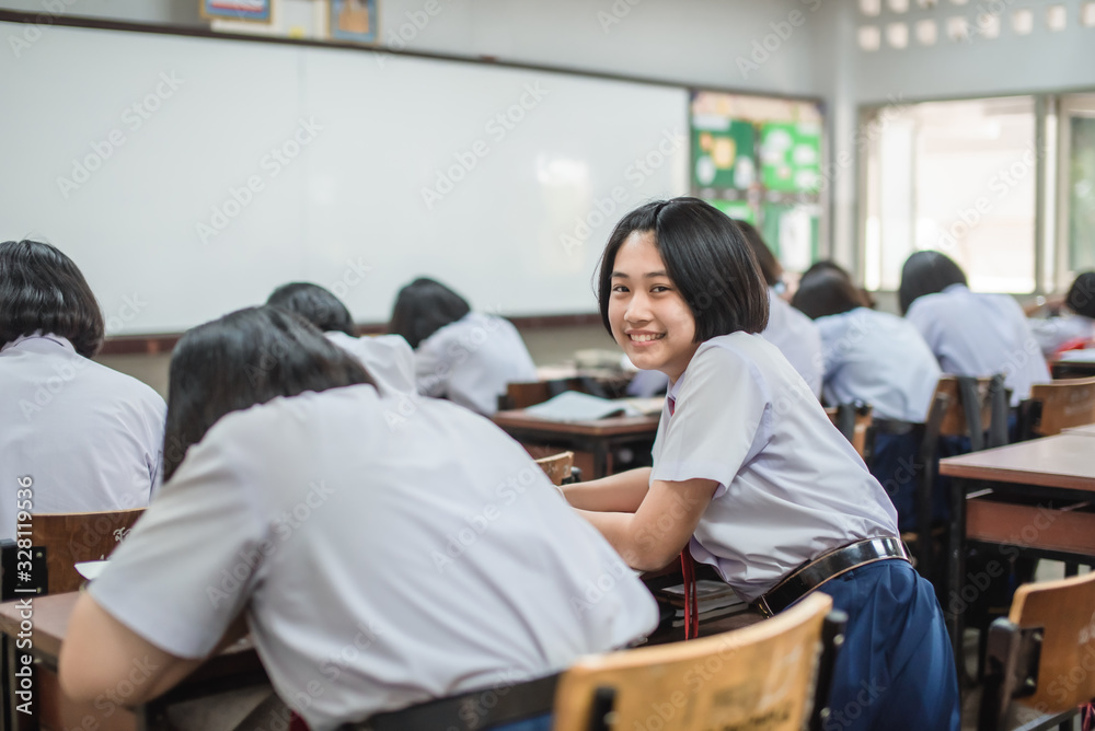 A pretty smiling Asian female student in white uniform is turning her face and smiling among her friends while studying in the classroom.