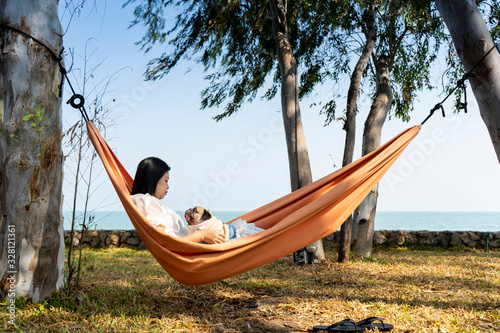 Funny cute pug dog smiling and tongue sticking out while on a trip with her owner  joyful young family  woman lying rest in comfortable hammock on sea beach. Happy times concept.