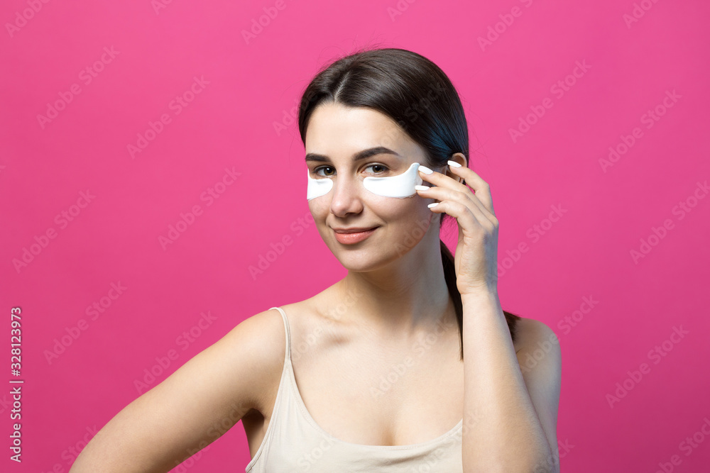 Close up portrait of pretty attractive girl with naked shoulders using patches under eyes. Standing over pink background. Close up portrait of pretty attractive girl with naked shoulders using patches under eyes. Standing over pink background.