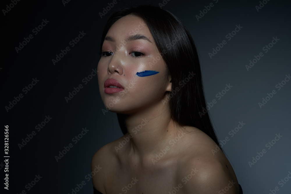 Portrait of a young beautiful Asian girl with smeared with oil blue paint, cheek in the form of a horizontal strip. The look leads away. On a gray background, a place to copy. The concept of creativ Portrait of a young beautiful Asian girl with smeared with oil blue paint, cheek in the form of a horizontal strip. The look leads away. On a gray background, a place to copy. The concept of creativ