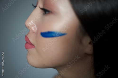 Profile of a young beautiful Asian girl with a cheek smeared with blue oil paint in the form of a horizontal stripe. Close-up photo. On a gray background  a place to copy