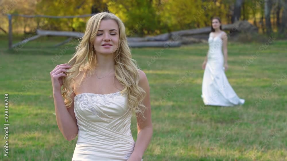 Beautiful blonde bride is posing at sunshine on the background brunette girl in wedding dress against autumn landscape Beautiful blonde bride is posing at sunshine on the background brunette girl in wedding dress against autumn landscape