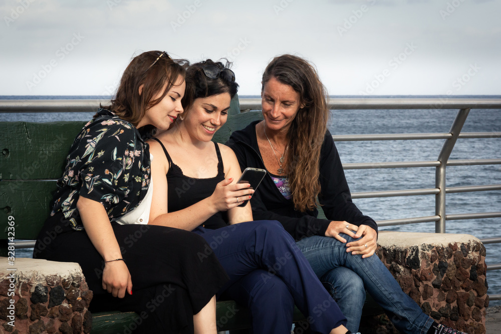 Group of girl friends having a good time sharing smart phone while sitting on bench by the ocean. Happy women using mobile outdoors