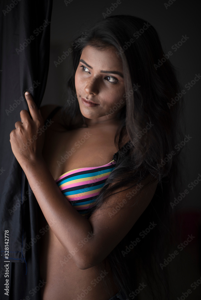 Portrait of young and beautiful dark skinned Indian Bengali woman in  colorful lingerie/bikini and hot pants standing in front of window and  looking outside in studio background. Boudoir photography. Stock Photo