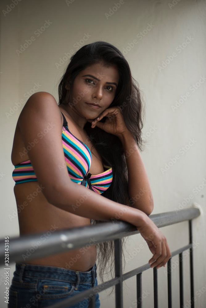 Stockfoto med beskrivningen Portrait of an young and beautiful dark skinned  Indian Bengali woman in colorful lingerie/bikini and hot pants posing in  casual mood on a balcony in white urban background. Boudoir