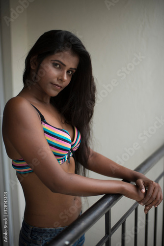 Portrait of an young and beautiful dark skinned Indian Bengali woman in colorful lingerie/bikini and hot pants posing in casual mood on a balcony in white urban background. Boudoir photography.