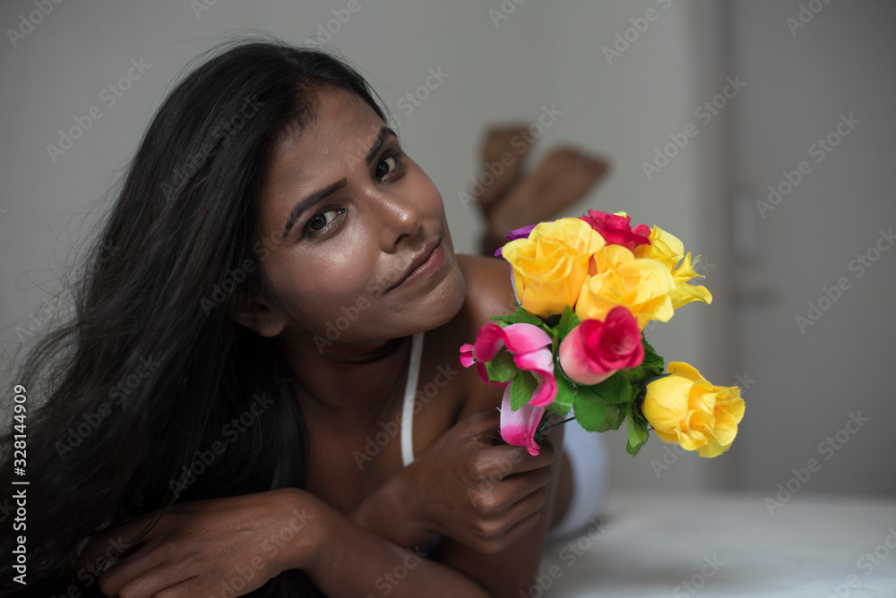 Portrait of an young dark skinned Indian Bengali woman in lingerie and vibrant flowers lying on  white bed in casual mood in white background. Indian lifestyle and  boudoir photography.