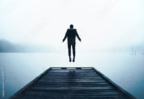 Man Jumping in Air Levitating at End of Wooden Dock Over Big Lake with Thick Layer of Fog and Clouds at Sunrise on Cold Brisk Wet Morning   photo